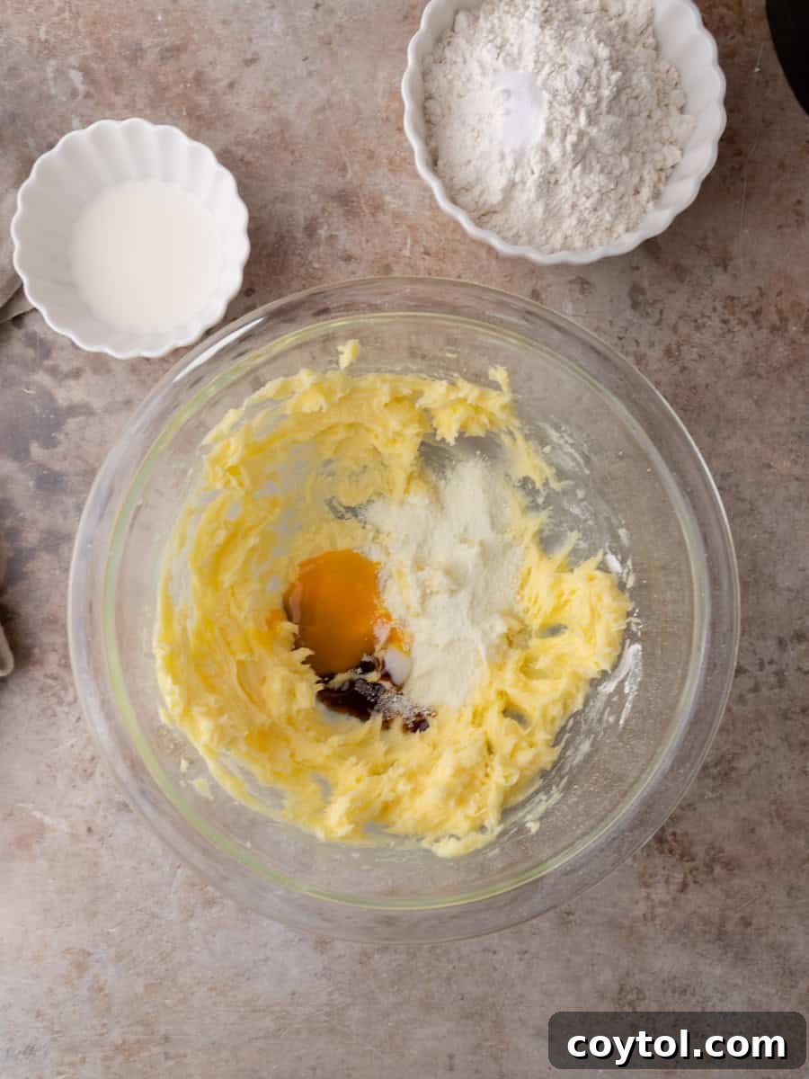 Softened butter, egg yolk, milk powder, and vanilla bean paste being mixed in a clear glass bowl, forming the creamy base for the cookie dough.