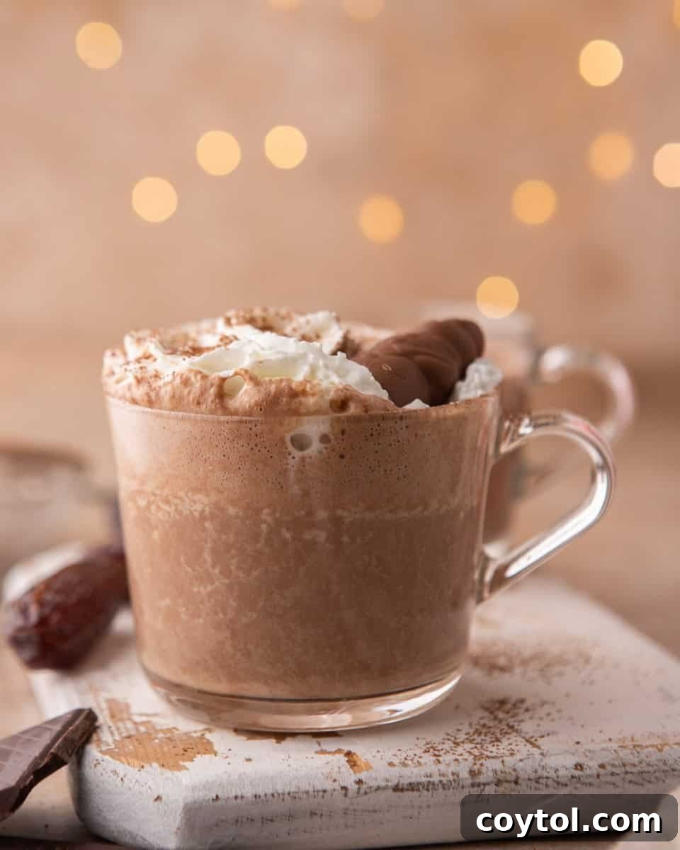 A glass mug filled with creamy date sweetened hot chocolate, generously topped with whipped cream and a sprinkle of chocolate shavings. The scene evokes warmth and indulgence.