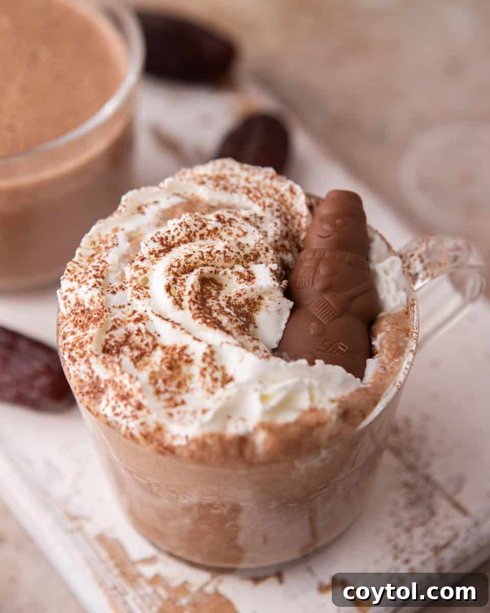 Close-up of a glass mug of date sweetened hot chocolate, lavishly topped with whipped cream and chocolate shavings, placed on a dark surface.
