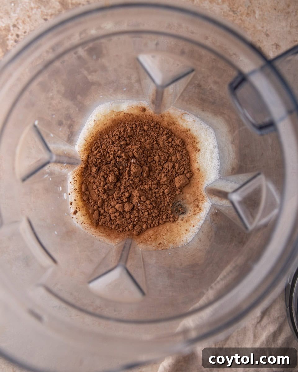 Cocoa powder being poured into a blender pitcher filled with milk, alongside Medjool dates.