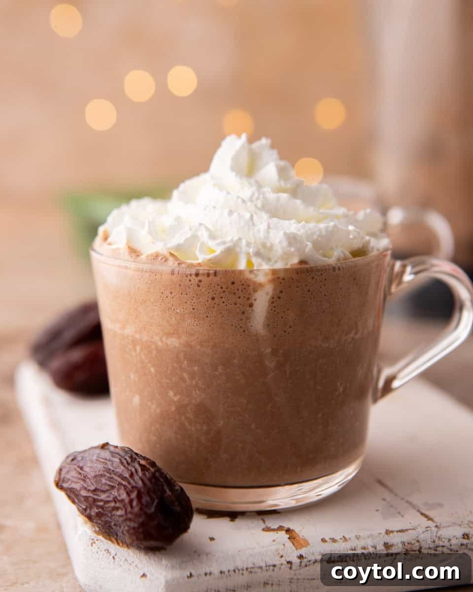 A clear glass mug showcasing the rich, creamy texture of date sweetened hot chocolate, topped generously with whipped cream and a light sprinkle of chocolate.