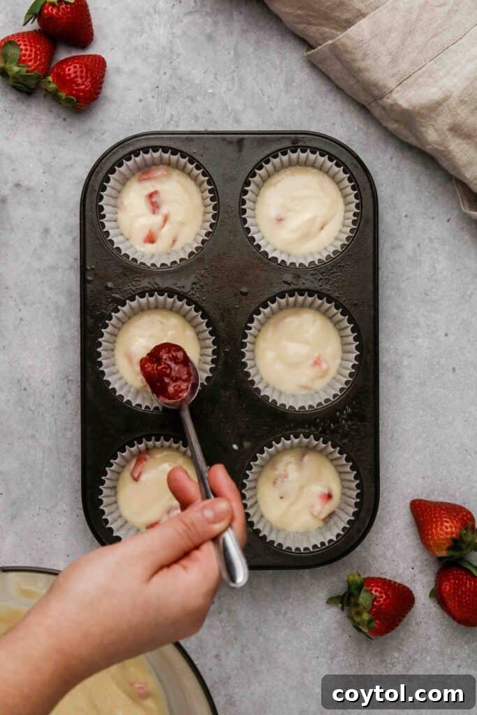 A spoonful of strawberry jam carefully placed in the center of a muffin cup filled with batter.