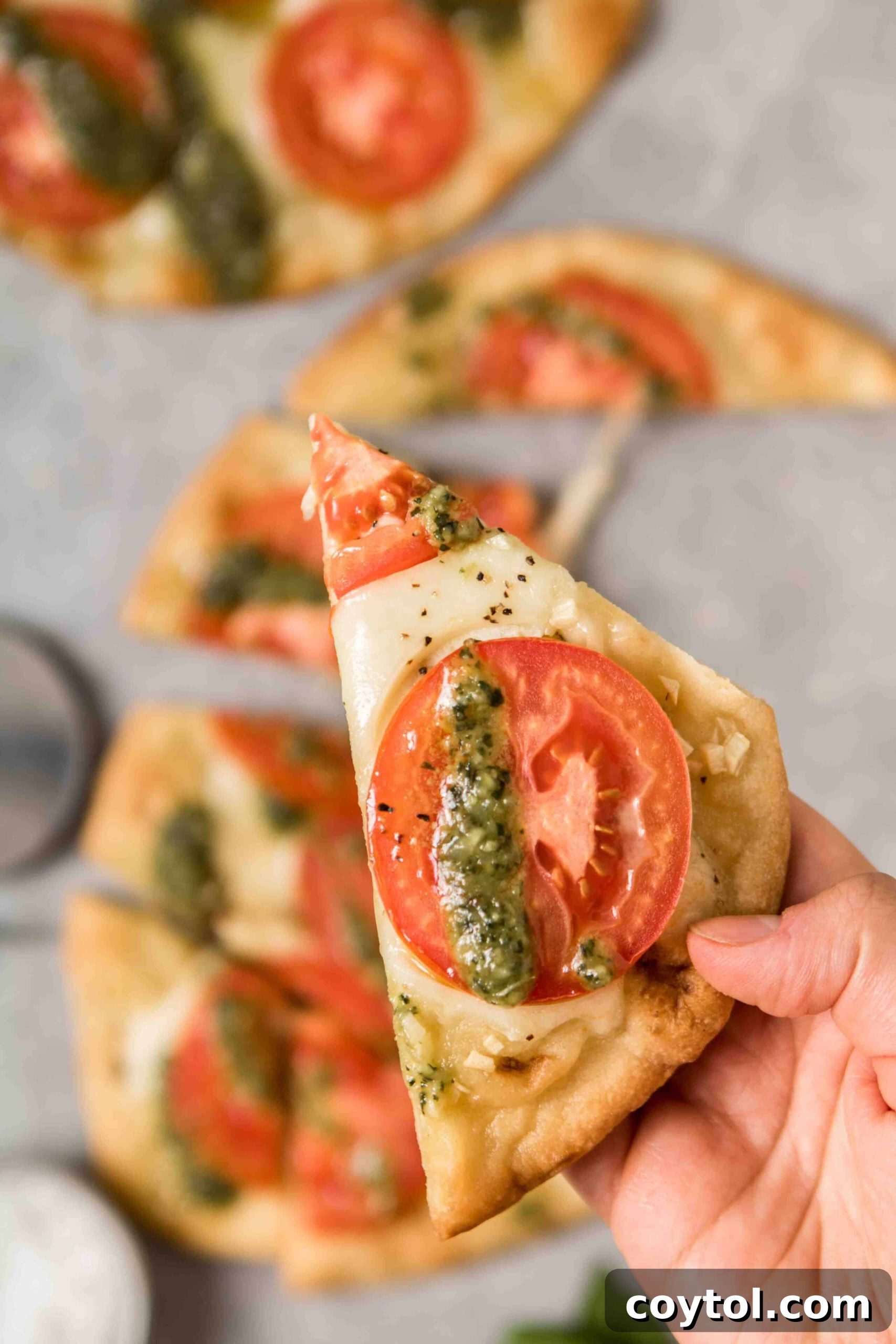 A hand holding a perfectly baked slice of naan bread pizza, adorned with vibrant pesto and melted cheese.