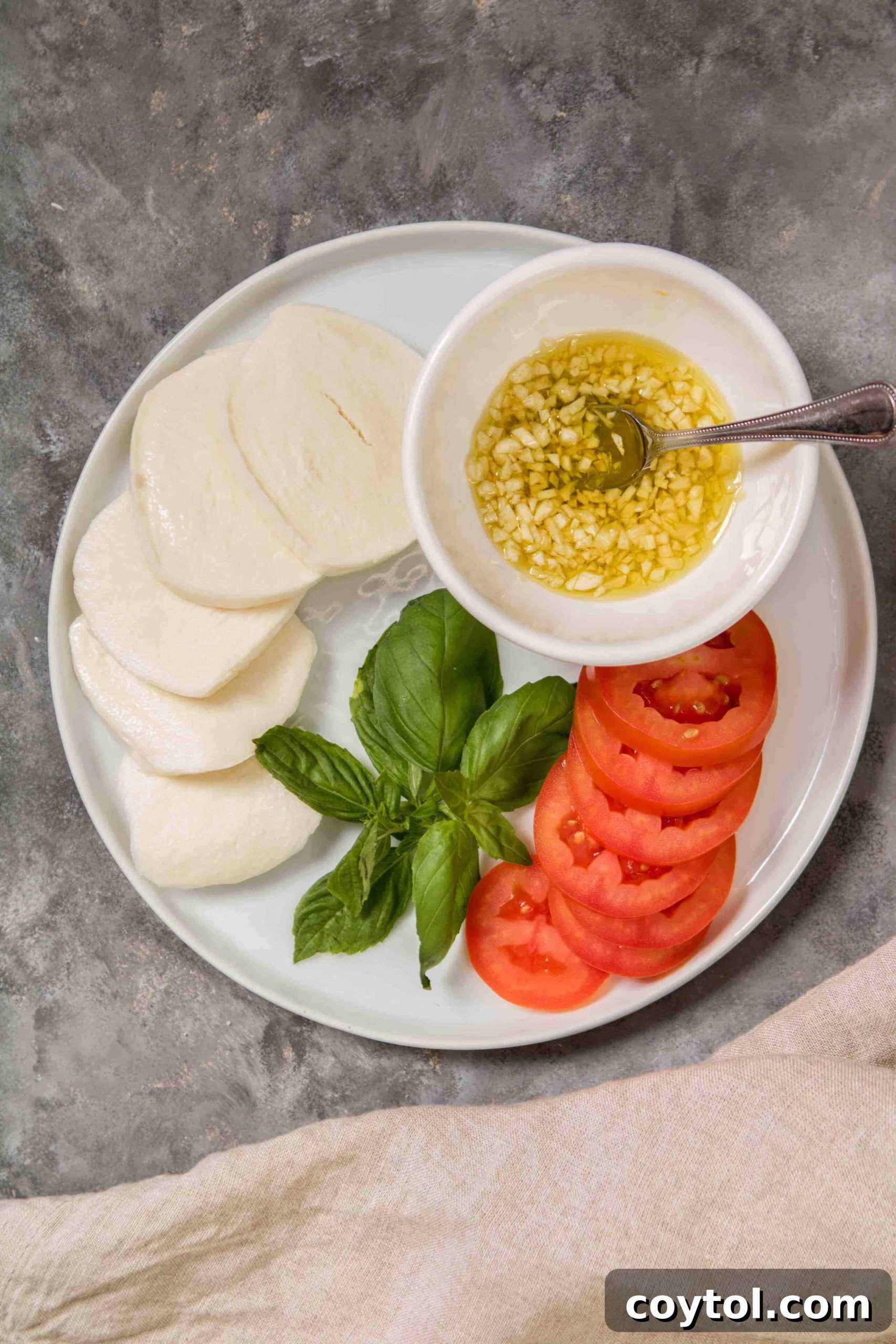 Arrangement of fresh ingredients for pesto pizza: vibrant basil leaves, slices of mozzarella, ripe Roma tomato slices, and minced garlic in olive oil.