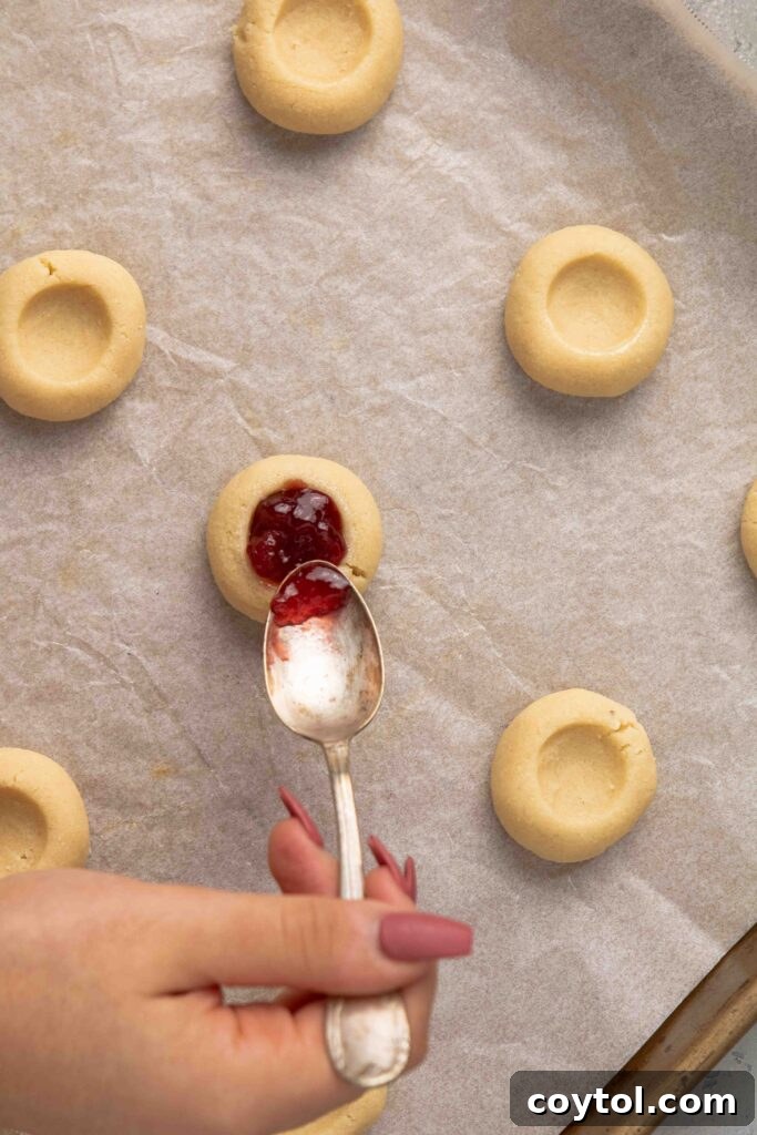Step-by-step image: Carefully filling the thumbprint cookies with jam using a small spoon before they are baked.
