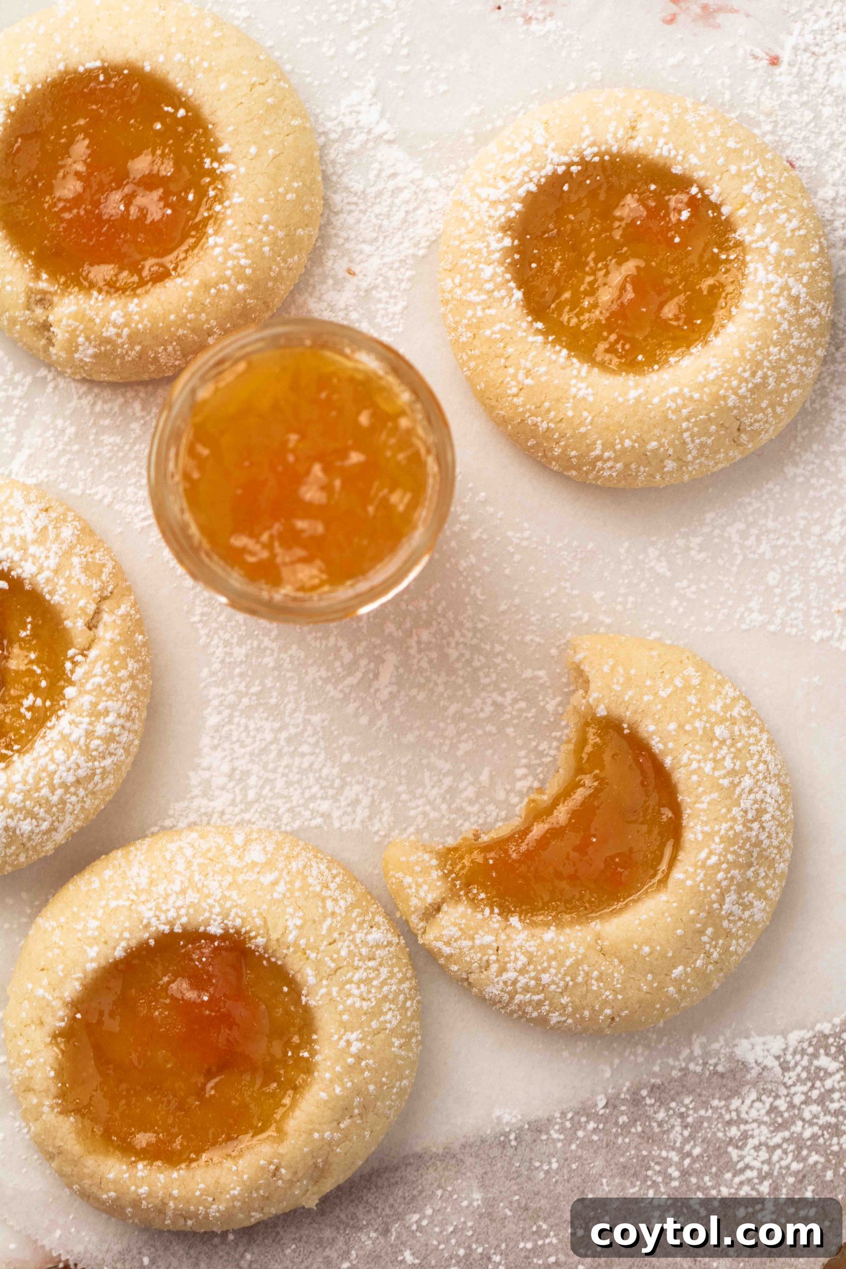 Overhead shot of apricot jam in a small glass bowl, surrounded by freshly baked golden thumbprint cookies with apricot filling, on a rustic wooden surface.