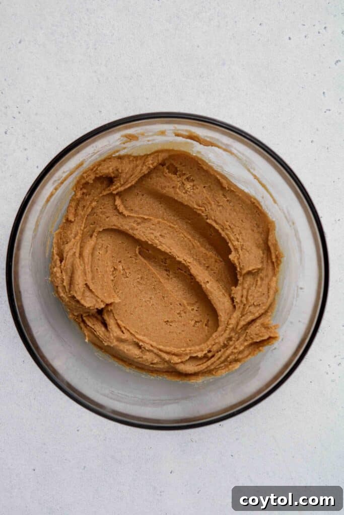 A close-up of a mixing bowl, showing peanut butter, sugar, and an egg being combined to form cookie dough.