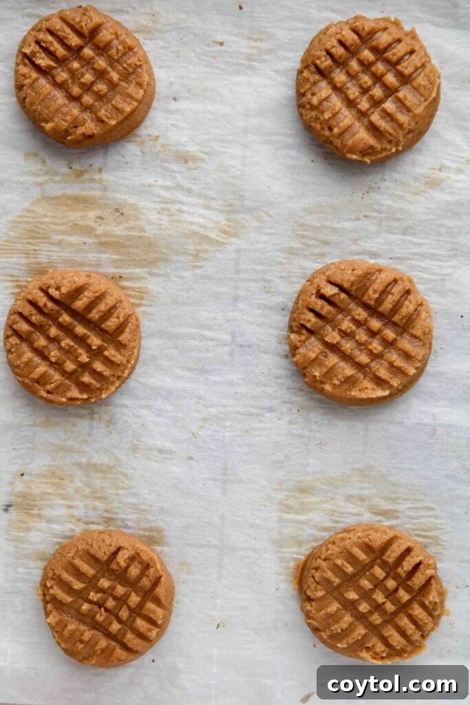 Hand pressing a fork into a scooped ball of peanut butter cookie dough to create the classic criss-cross pattern before baking.
