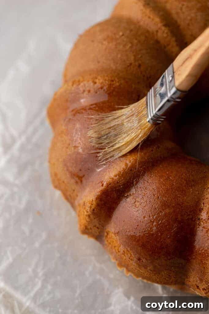 A warm, freshly baked bundt cake being meticulously brushed with melted butter, preparing its surface for the crucial cinnamon-sugar coating that defines its apple cider donut flavor.