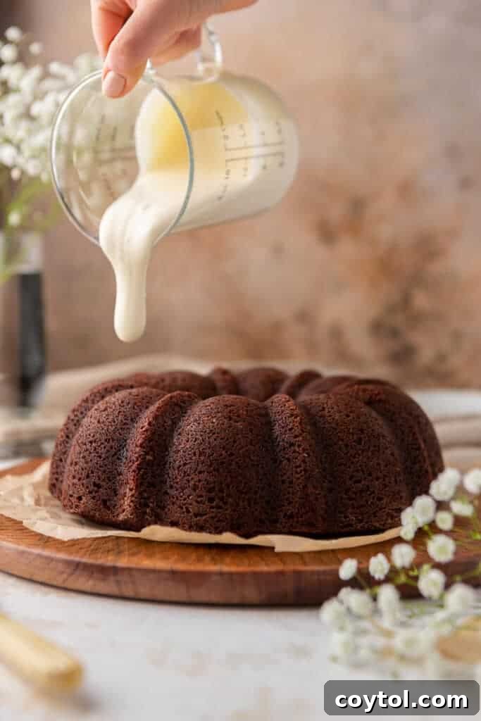 A hand is gently pouring a smooth cream cheese glaze over a beautifully baked red velvet bundt cake, highlighting its rich color.
