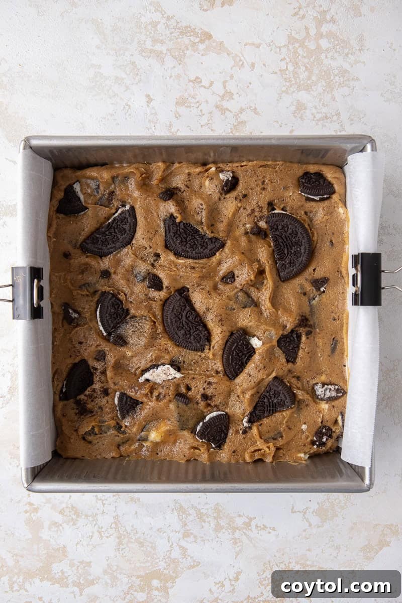 Unbaked Oreo blondie batter spread evenly in a parchment-lined 8x8 inch baking pan, with extra chopped Oreos sprinkled on top.