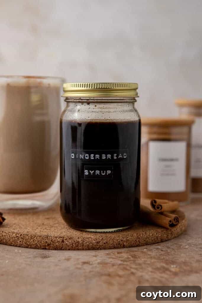A styled photo of gingerbread syrup in a jar with festive background elements like cinnamon sticks and a latte to show serving inspiration.