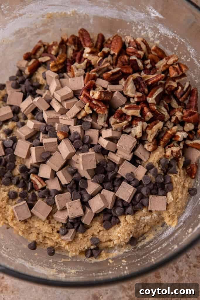Hands adding a generous amount of chocolate chunks and chopped toasted pecans to a bowl of freshly made cookie dough, illustrating the mix-in step.