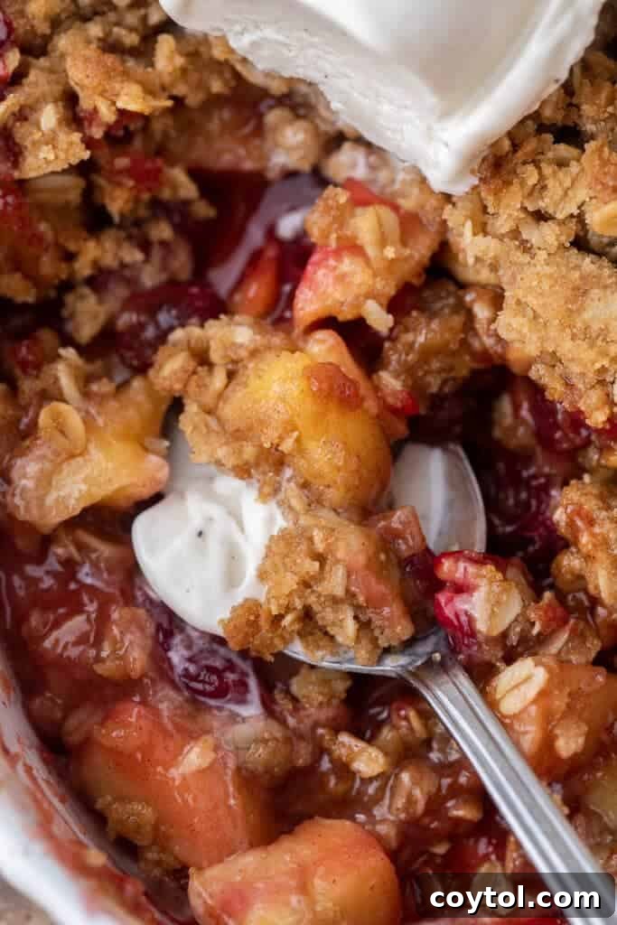 A spoon digging into a warm cranberry apple crisp with a generous scoop of vanilla ice cream on top, creating a perfect bite.