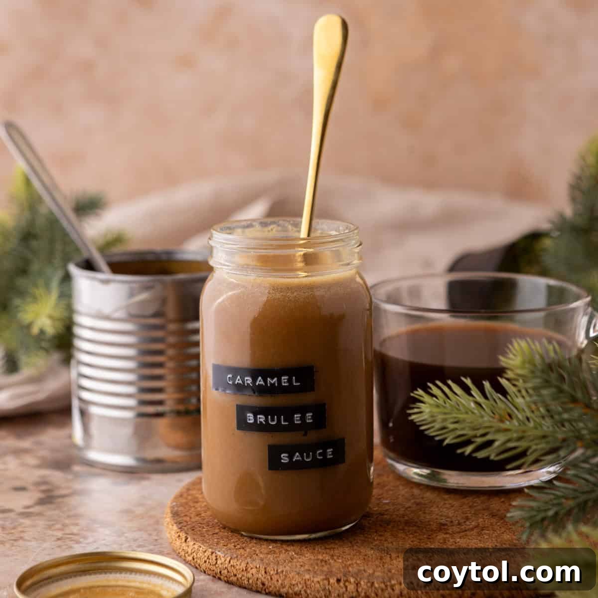 Jar of homemade Starbucks Caramel Brulee Sauce with gold spoon, coffee cup, and festive greenery on a cozy kitchen counter.