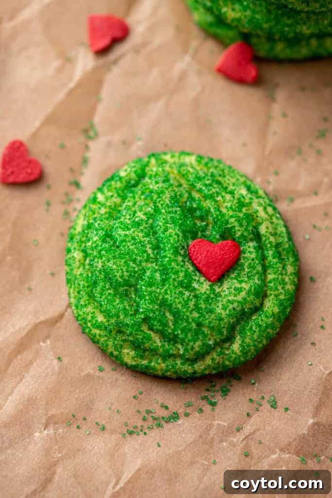 Grinch sugar cookies, vibrant green and topped with tiny red hearts, arranged on a baking sheet, ready to be enjoyed during the holiday season.