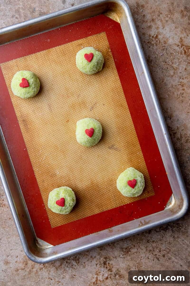 Grinch sugar cookie dough balls with red heart sprinkles on a baking sheet, perfectly spaced for even baking.