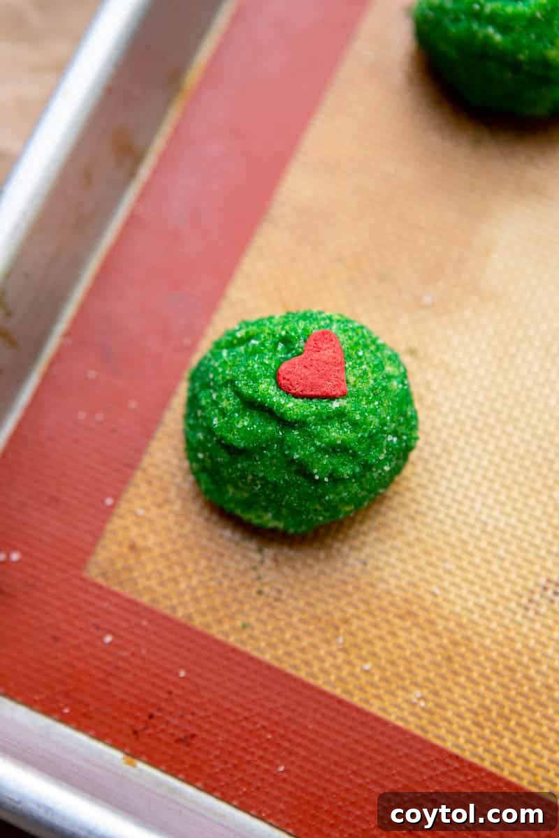 Close-up of unbaked Grinch sugar cookie dough balls adorned with red heart sprinkles on parchment paper.