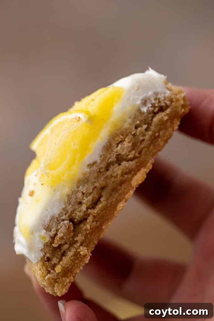 Divine Crumbl Lemon Cheesecake Cookies 12 A hand holding a sliced lemon cheesecake cookie, revealing its perfectly layered interior with the cookie base, frosting, and lemon curd.