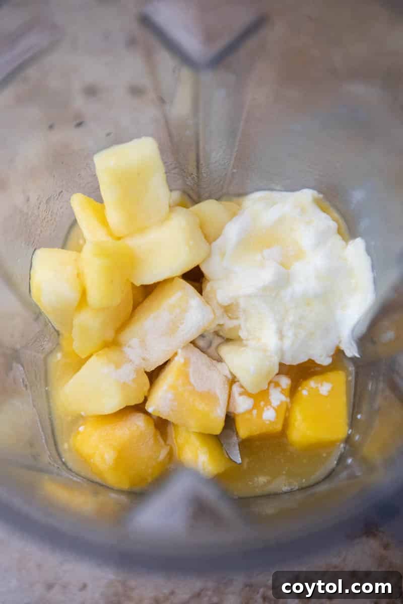 Frozen mango and pineapple chunks along with liquids being added to a blender jar.