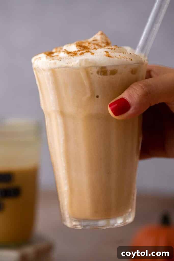Hand holding a tall glass of Starbucks Pumpkin Spice Frappuccino with whipped cream outdoors.