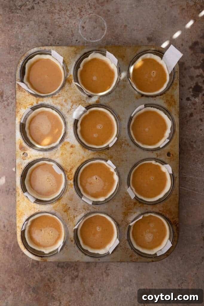 Raw pie dough pressed into individual muffin tin cups, filled with pumpkin batter, before baking.