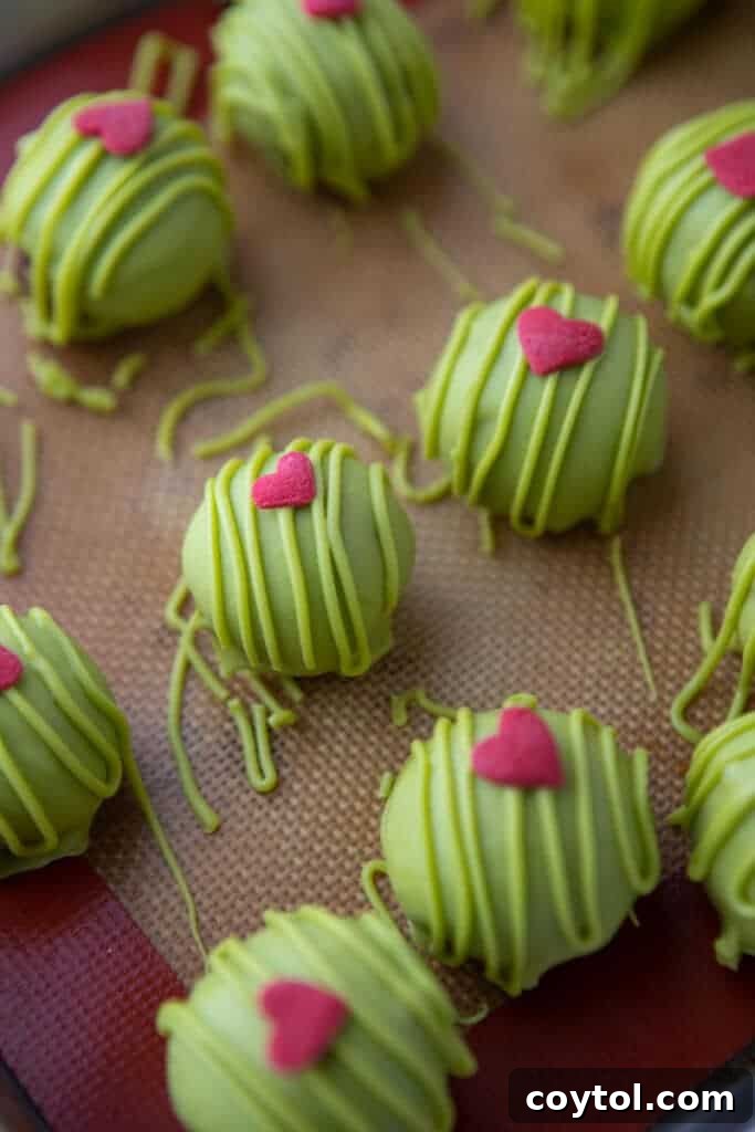 a bunch of rolled out green coated grinch oreo truffles laid out on a baking sheet, with red hearts