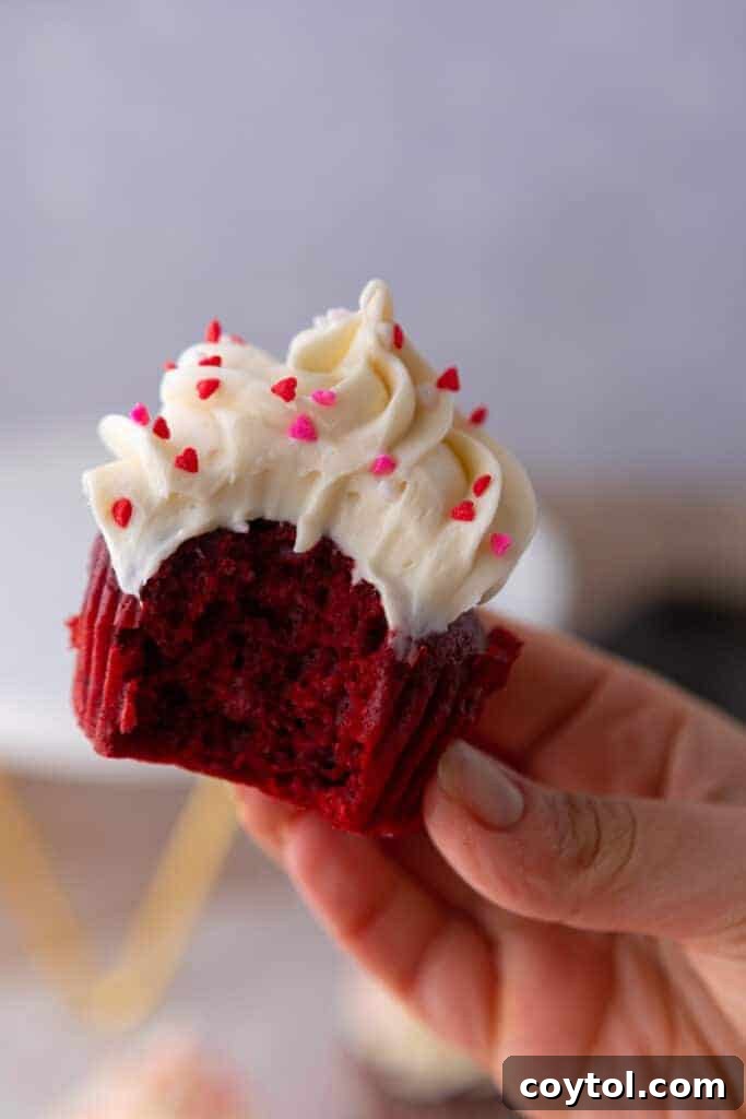 A hand delicately holds one of the small batch Red Velvet cupcakes, revealing a perfect bite taken out, highlighting its moist interior and generous cream cheese frosting.