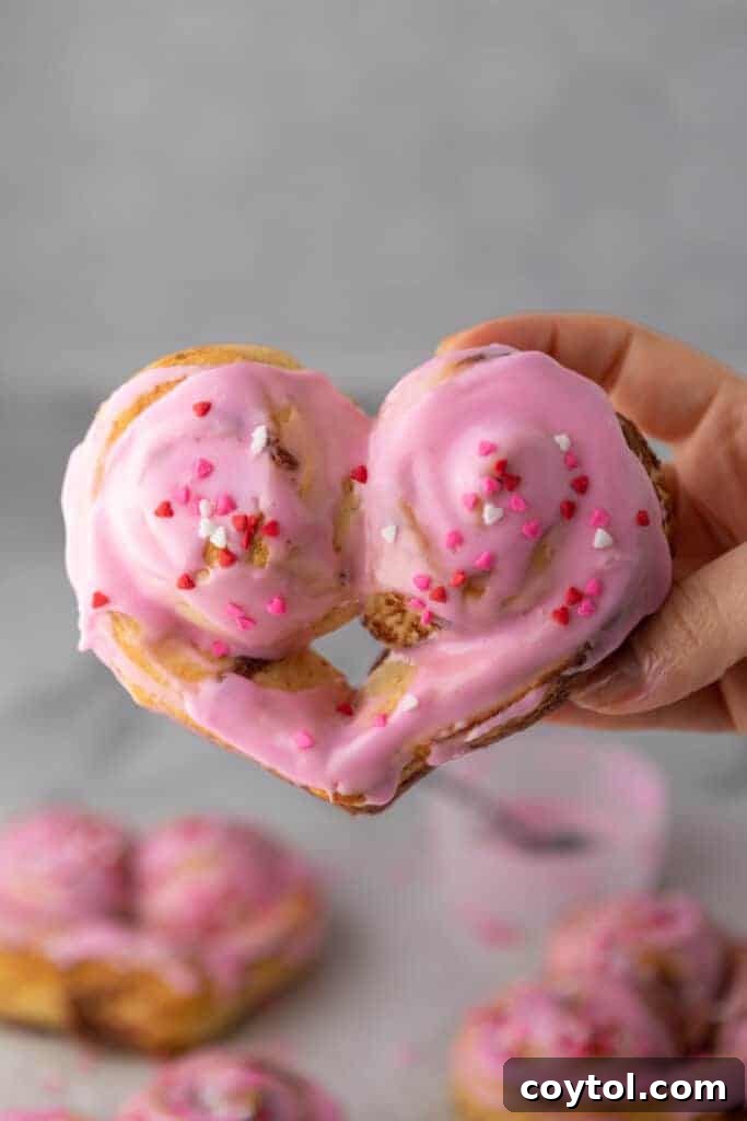 A hand holding a perfectly baked heart-shaped cinnamon roll, featuring a generous swirl of pink frosting and colorful Valentine’s sprinkles, against a softly blurred background.