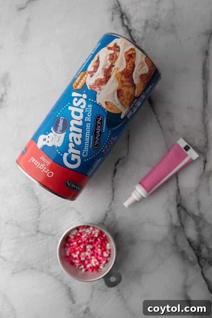 A minimalist shot of the ingredients laid out on a clean kitchen counter, including a can of cinnamon rolls, a small bottle of pink food coloring, and a container of festive Valentine’s sprinkles.