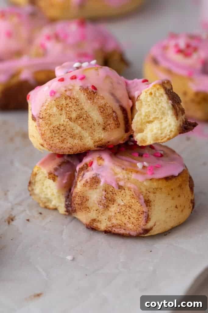 A stack of three freshly baked heart-shaped cinnamon rolls, each generously covered in vibrant pink frosting and adorned with colorful Valentine’s sprinkles, showcasing their soft, inviting centers.
