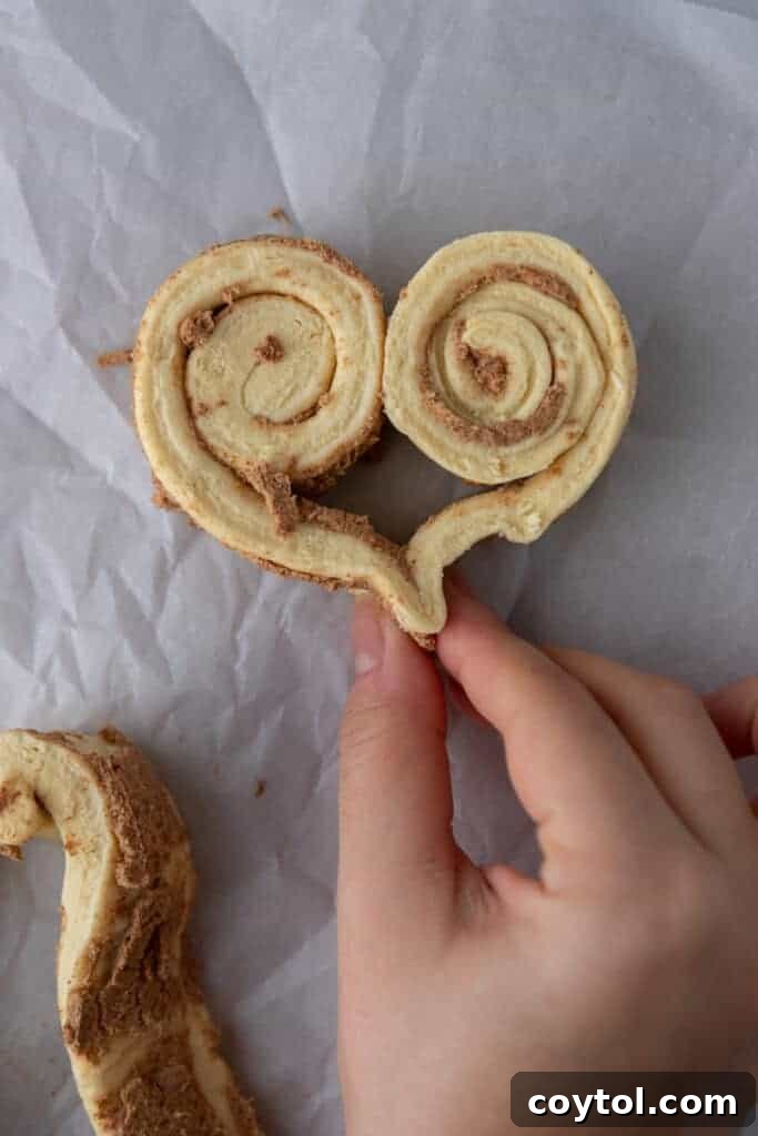 A close-up shot of hands gently molding and pinching cinnamon roll dough into a heart shape, illustrating the delicate process of crafting each individual roll.