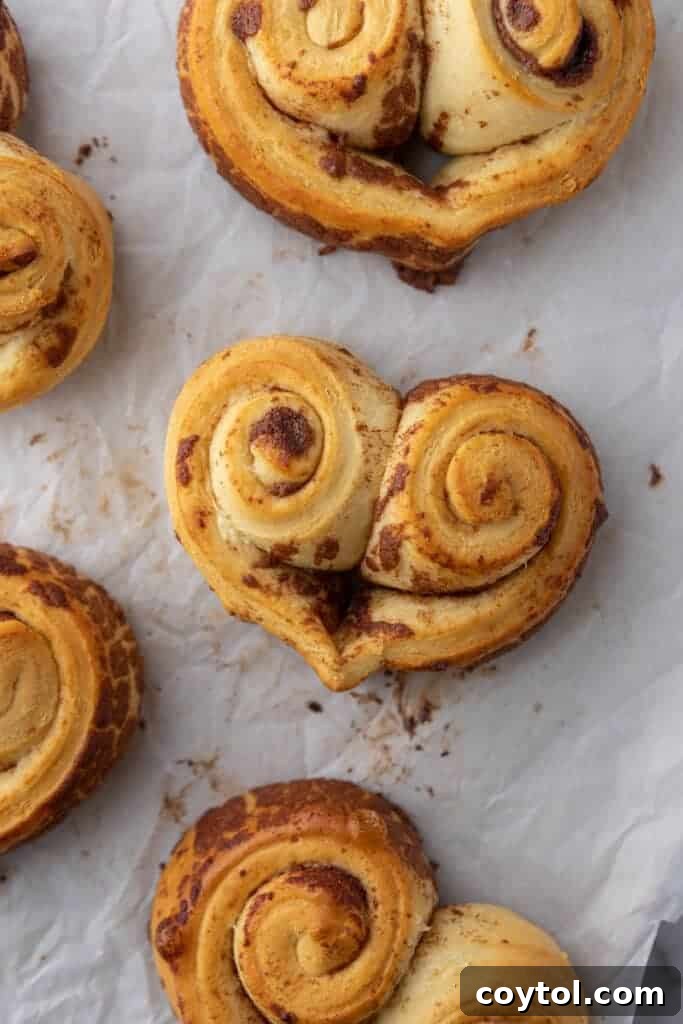 A baking sheet showcasing several baked heart-shaped cinnamon rolls, still on parchment paper, glowing golden brown and awaiting their sweet pink frosting and festive sprinkles.