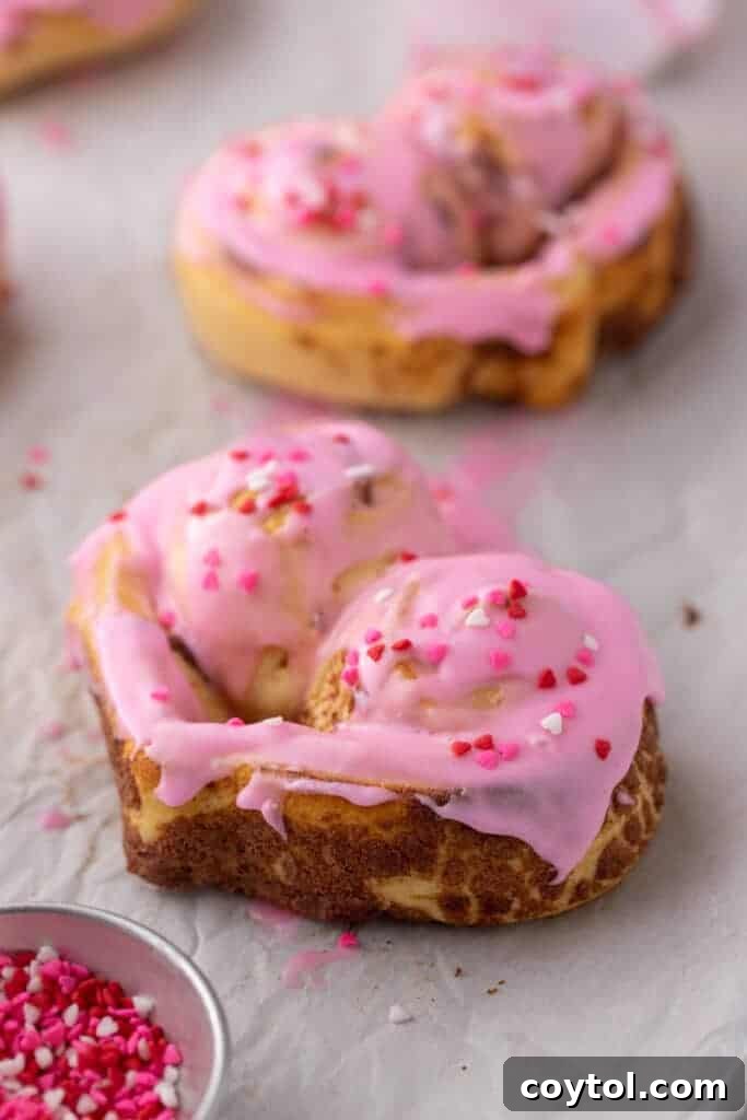A stunning close-up of a single heart-shaped cinnamon roll, beautifully frosted with pink icing and adorned with Valentine’s sprinkles, highlighting its irresistible texture and festive appeal.
