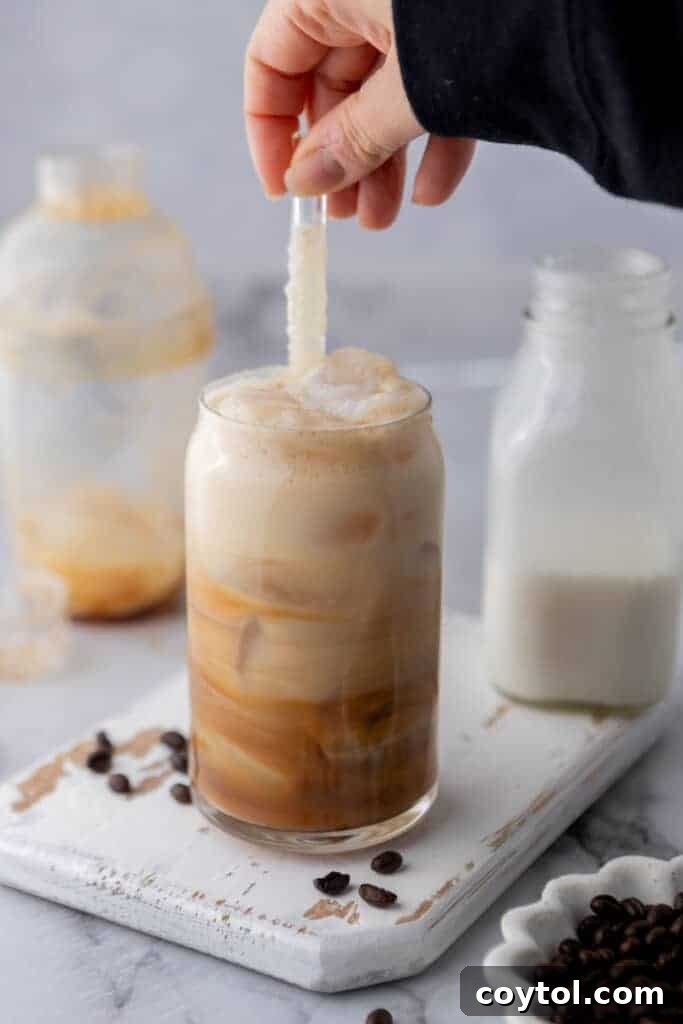 A hand gently stirring an Iced Hazelnut Oatmilk Shaken Espresso with a straw, showcasing the blend of flavors.