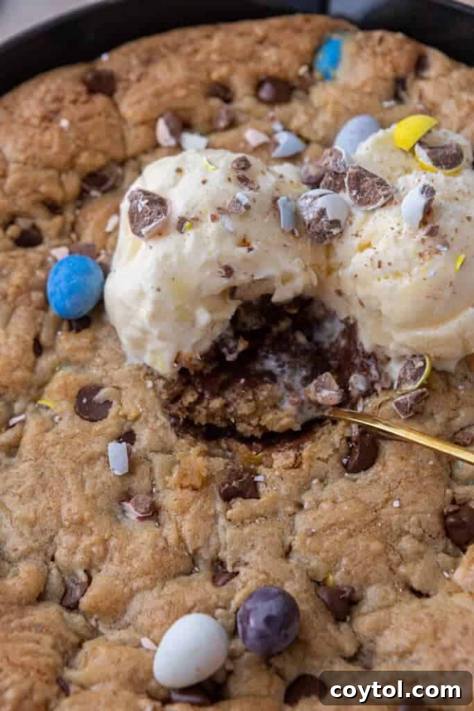 A close-up shot of a fork lifting a perfect bite of the Cadbury Mini Egg Skillet Cookie, showing the soft, gooey interior and melted chocolate, topped with a dollop of vanilla ice cream.