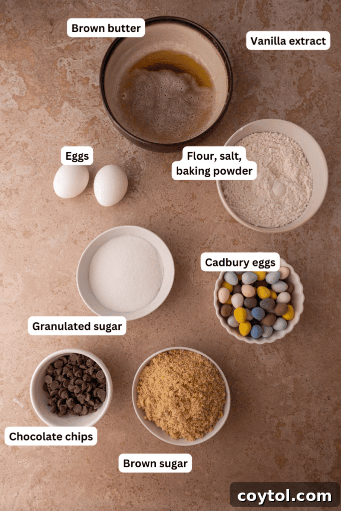An organized collection of baking ingredients on a wooden kitchen counter, including butter, various sugars, eggs, flour, and a bowl of colorful Cadbury Mini Eggs, all ready for baking.