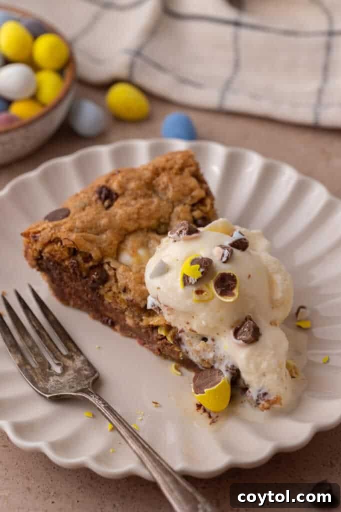 A perfectly portioned slice of Cadbury Mini Egg Skillet Cookie on a white plate, generously topped with a melting scoop of vanilla ice cream, with a fork poised for a bite.