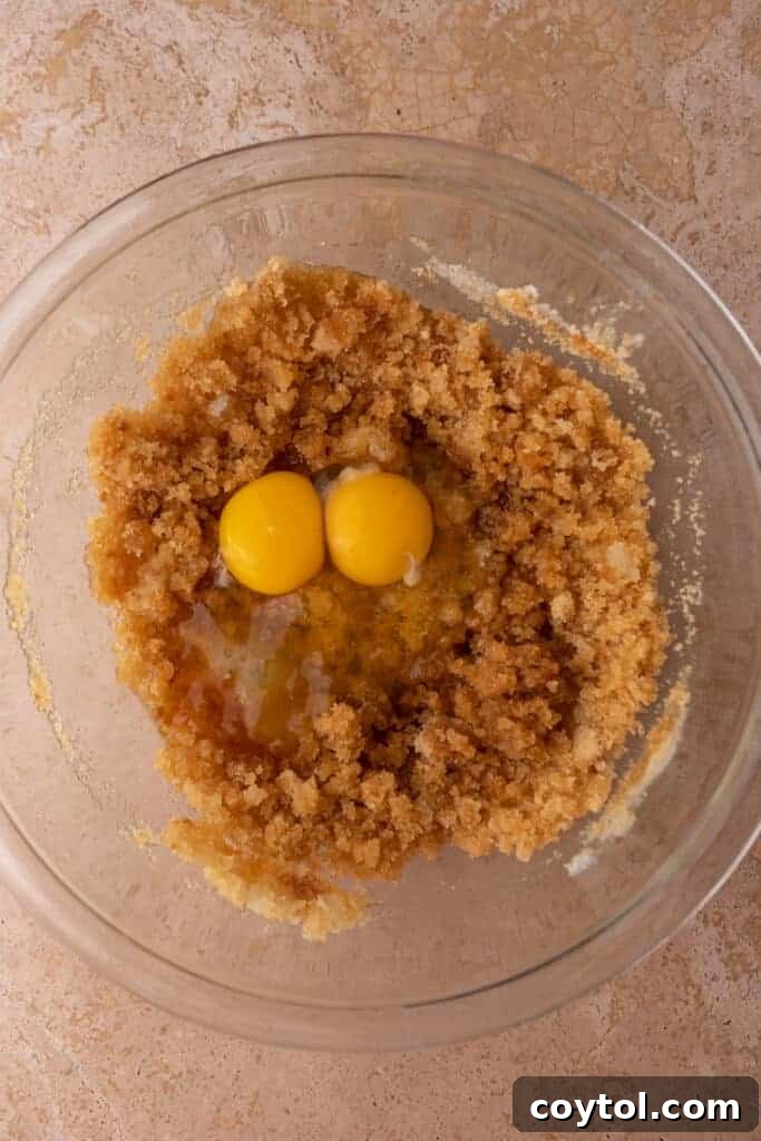 Two eggs being cracked and dropped into a bowl of creamy, light-colored cookie batter, illustrating the step of incorporating wet ingredients.