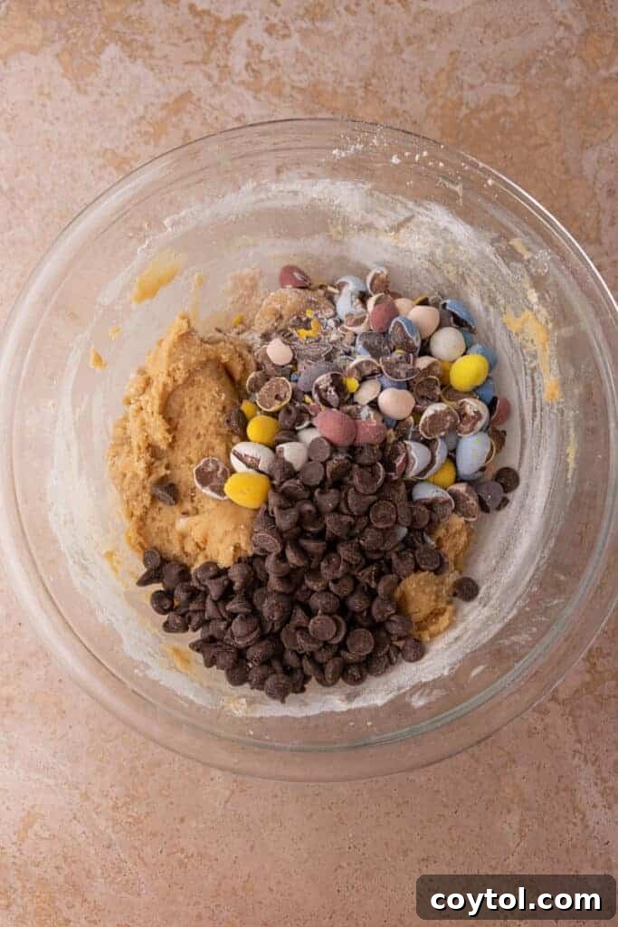 Colorful crushed Cadbury Mini Eggs and semi-sweet chocolate chips being gently folded into the mixed cookie dough in a large bowl, demonstrating the final ingredient incorporation step.