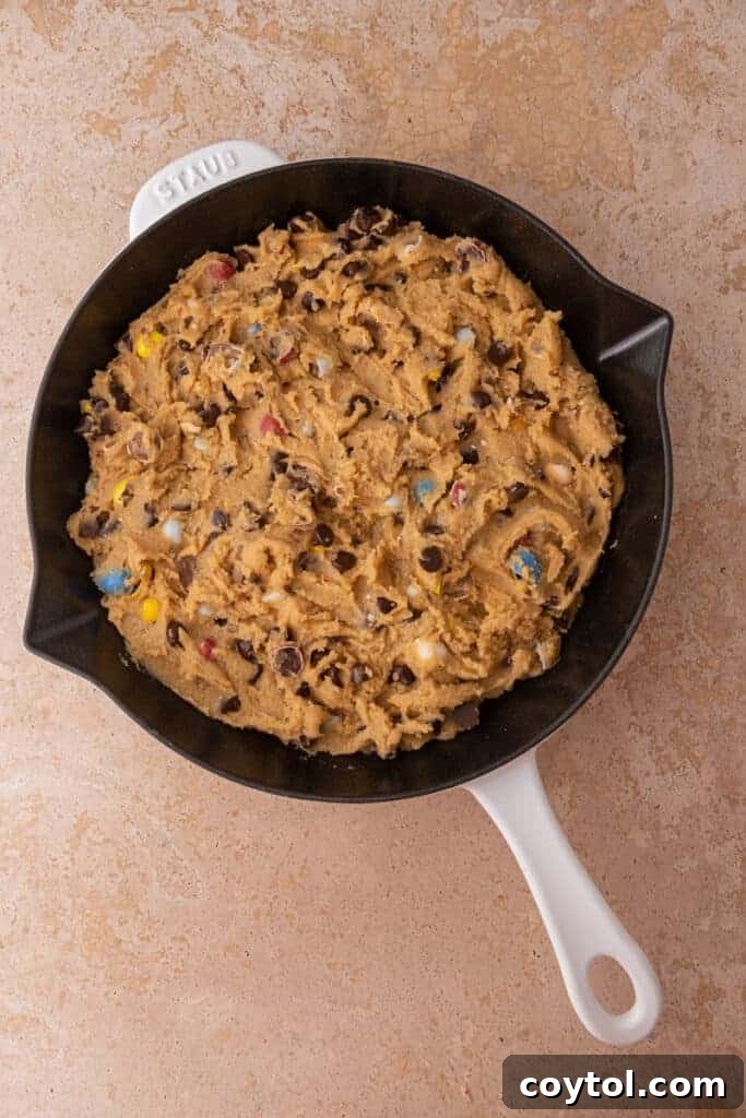 Raw cookie dough spread evenly across the bottom of a cast iron skillet, before being placed into the oven for baking.