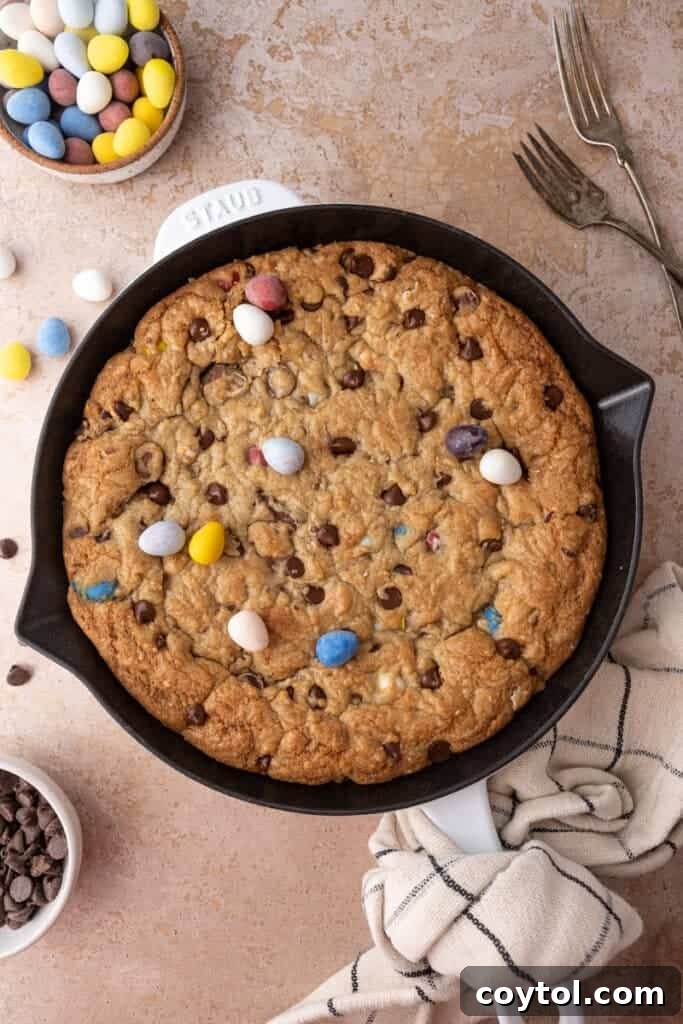 A fully baked Cadbury Mini Egg Skillet Cookie, golden brown with visible melted chocolate and colorful mini eggs, fresh from the oven in its cast iron skillet.