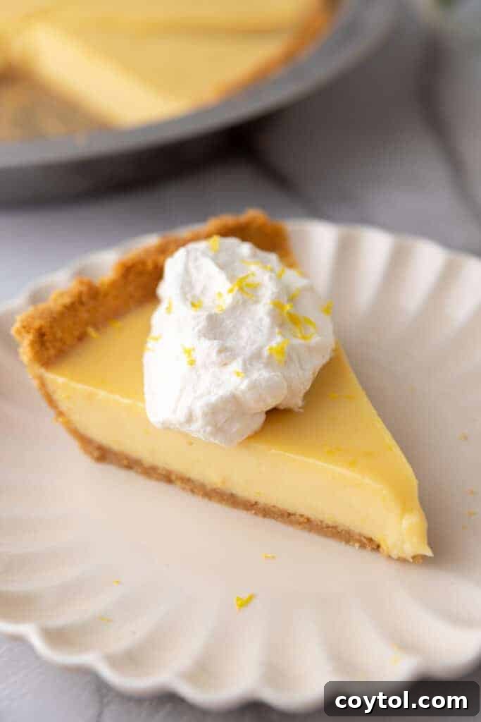 A neatly cut slice of lemon pie with graham cracker crust on a white plate, garnished with whipped cream and lemon zest.