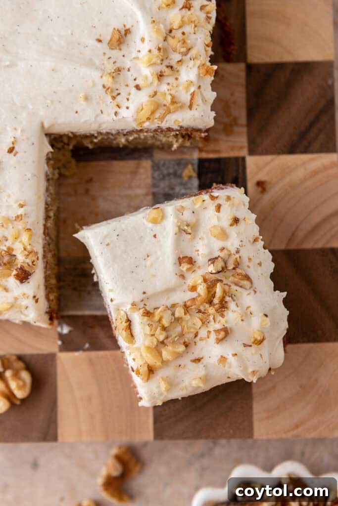 A serving spatula lifting a slice of frosted banana cake from the pan, showing the moist interior.