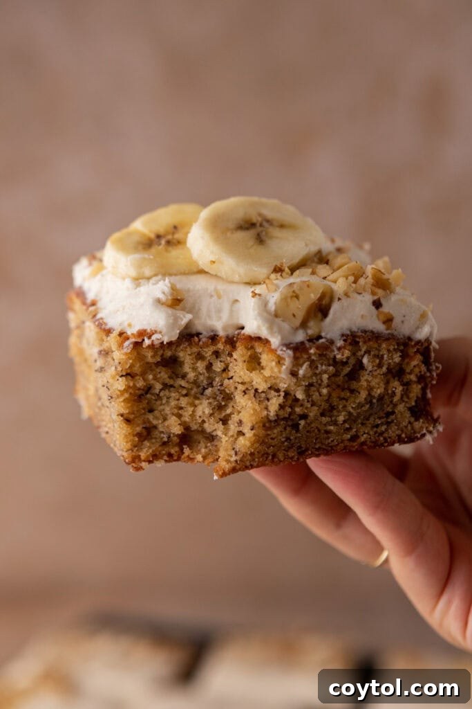 A hand holding up a slice of golden-brown banana cake topped with fluffy white cream cheese frosting.