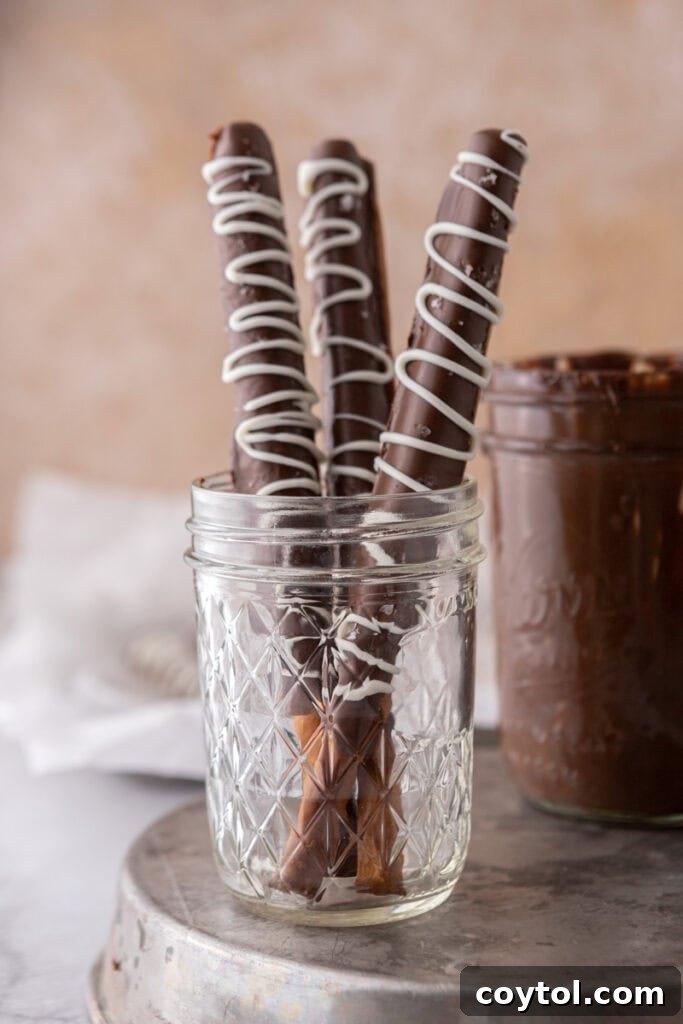 Three caramel chocolate covered pretzel rods, perfectly arranged in a festive cup, showcasing their golden caramel layer and rich chocolate coating, topped with a sprinkle of sea salt.