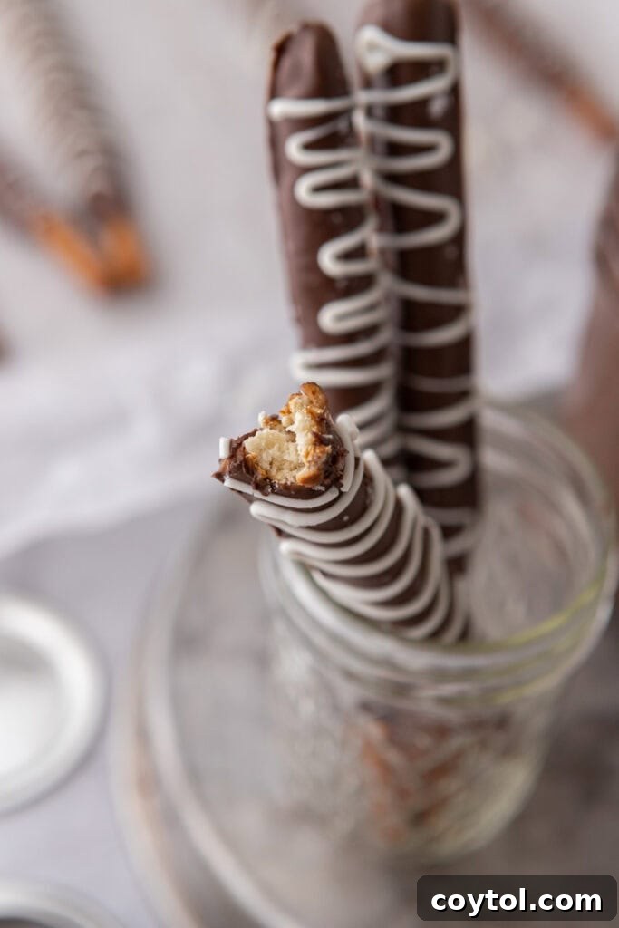 A close-up of three caramel chocolate covered pretzel rods in a glass cup, highlighting the textured toppings and rich layers, ready to be enjoyed.