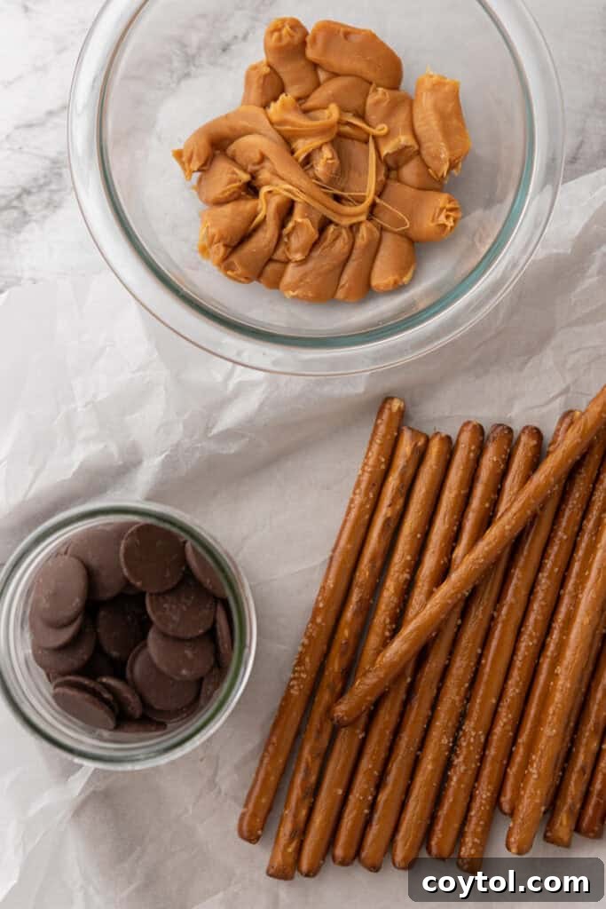 A curated selection of ingredients laid out on a clean surface, ready for making caramel chocolate covered pretzel rods, including pretzel rods, soft caramels, heavy cream, and chocolate melting wafers.