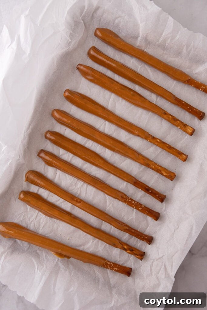 Caramel coated pretzel rods resting on a parchment-lined baking sheet, allowing the caramel to set.