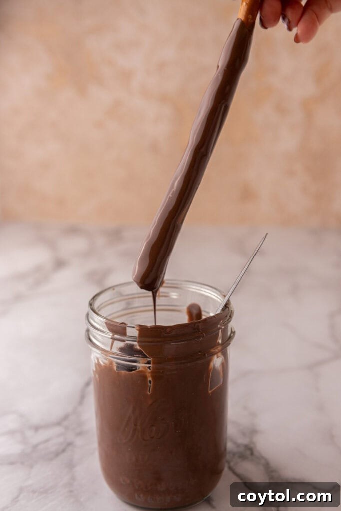 A caramel-coated pretzel rod being dipped into a bowl of melted chocolate, creating the second layer.